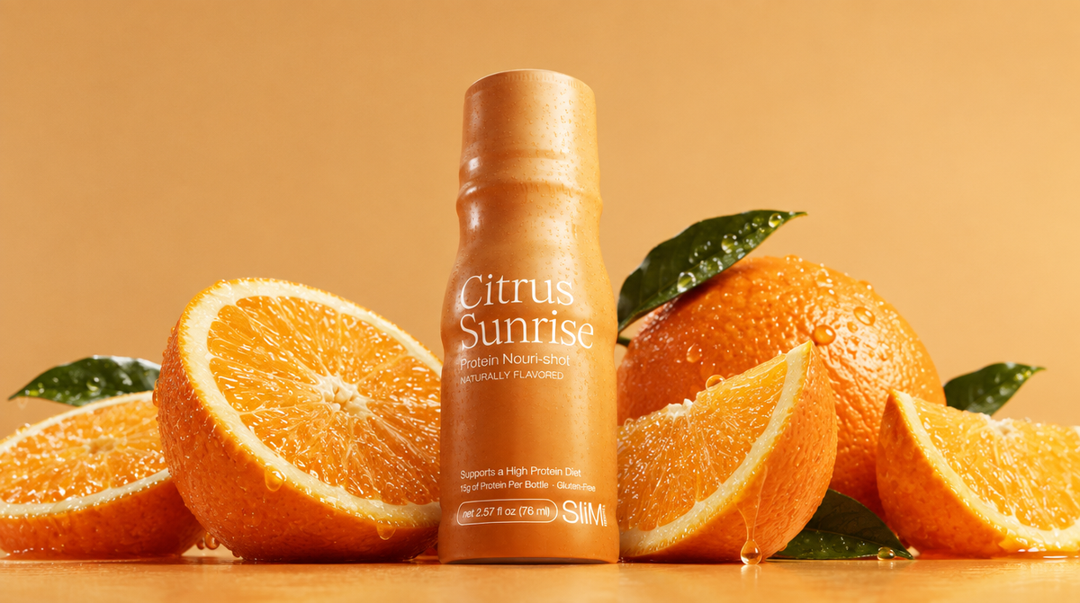 A bottle of SLIM Citrus Sunrise protein Nouri-Shot stands centered among fresh orange slices and whole oranges on a warm orange-brown background.

